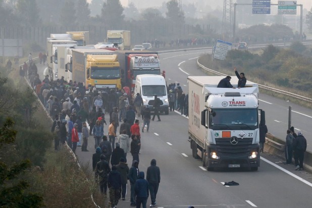 Migrants-gather-on-the-road-as-two-board-a-lorry-on-the-access-road-to-reach-the-ferry-terminal-in-Calais