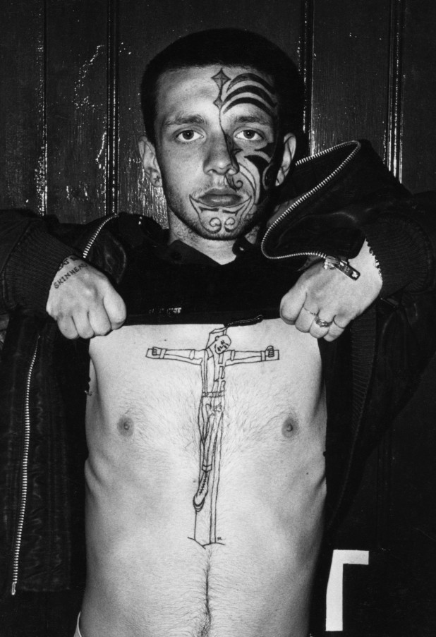 Skinhead with facial tattoo lifting up his top to reveal a 'skinhead on the cross' tattoo on his chest