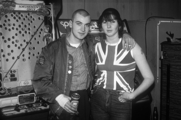 A skinhead couple wearing Union Jack T-shirt, flying jacket, Ben Sherman shirt, badges, cropped hair, tattoos, drinking beer and smoking at an arcade in Brighton, UK 1985.